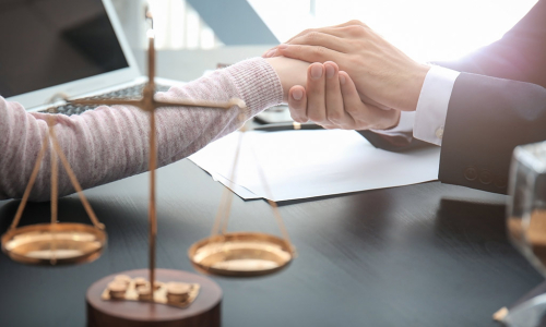 Hand eines Anwalts hält Hand einer Klientin. Im Vordergrund die Waage der Justitia.