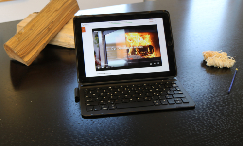 Ein Tablet auf einem Tisch, daneben und im Hintergrund ein Kaminanzünder und ein Holzscheit. Auf dem Display sieht man ein Video über das Feuermachen im Kaminofen
