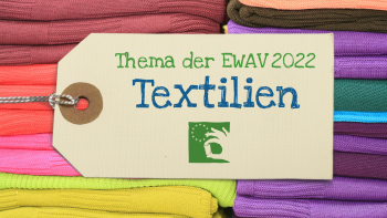 gestapelte Textilien, davor großes Etikett mit Aufschrift "Thema der EWAV 2022 Textilien"